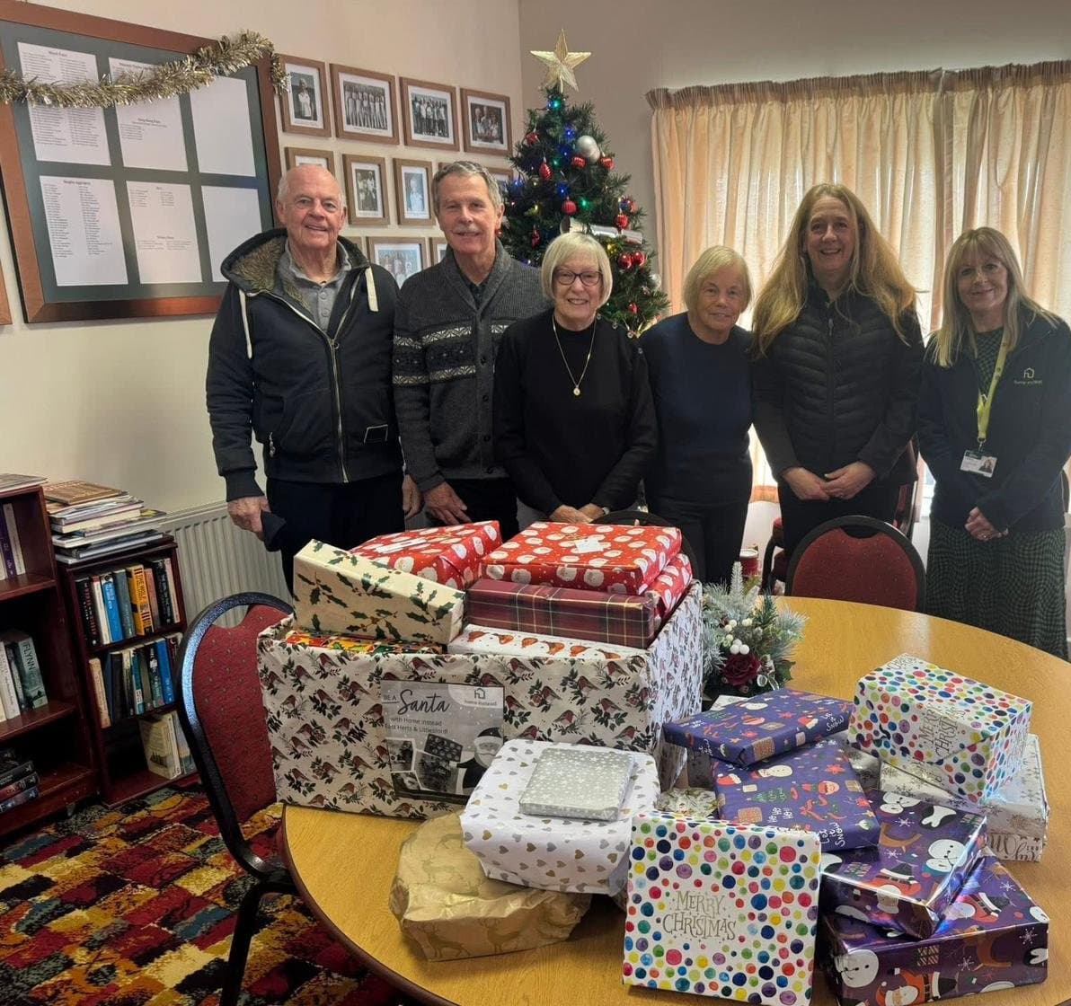 Six people stand behind a table with wrapped Christmas presents in a festive, decorated room with a tree. - Home Instead