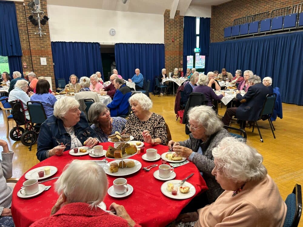Elderly people enjoying tea and cake together at round tables in a community hall. - Home Instead