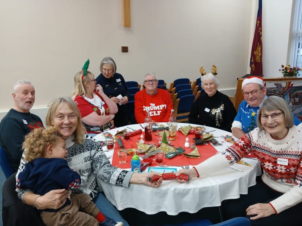 A group of people in festive attire sit smiling around a decorated table at a holiday gathering. - Home Instead