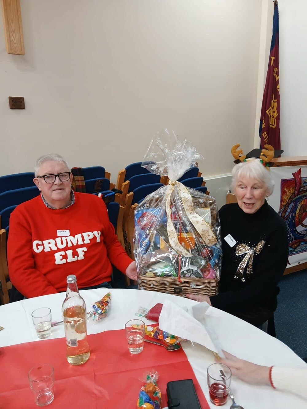 Two seniors sit at a table, smiling, holding a gift basket; one wears a "Grumpy Elf" sweater. - Home Instead