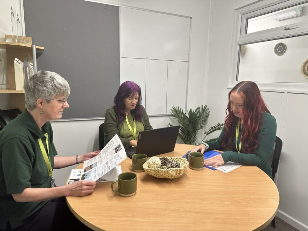 Three women sit at a round table with mugs, reading documents and working on a laptop in an office. - Home Instead