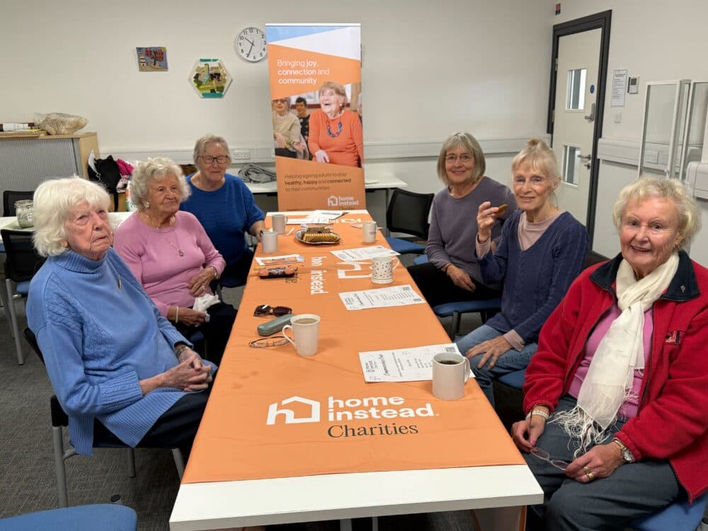 Six older women sit around a table with tea and snacks at a "Home Instead Charities" community event. - Home Instead