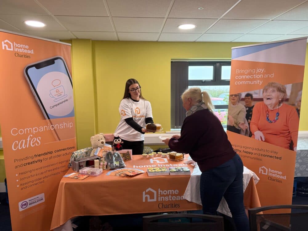 Two women interact at a Home Instead Charities table with gifts, banners, and information about companionship cafés. - Home Instead