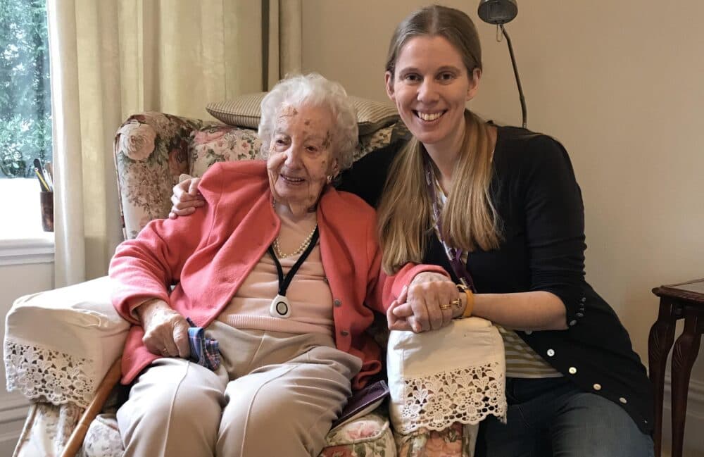 An elderly woman and a younger woman smile while sitting together on a floral armchair in a cozy room. - Home Instead