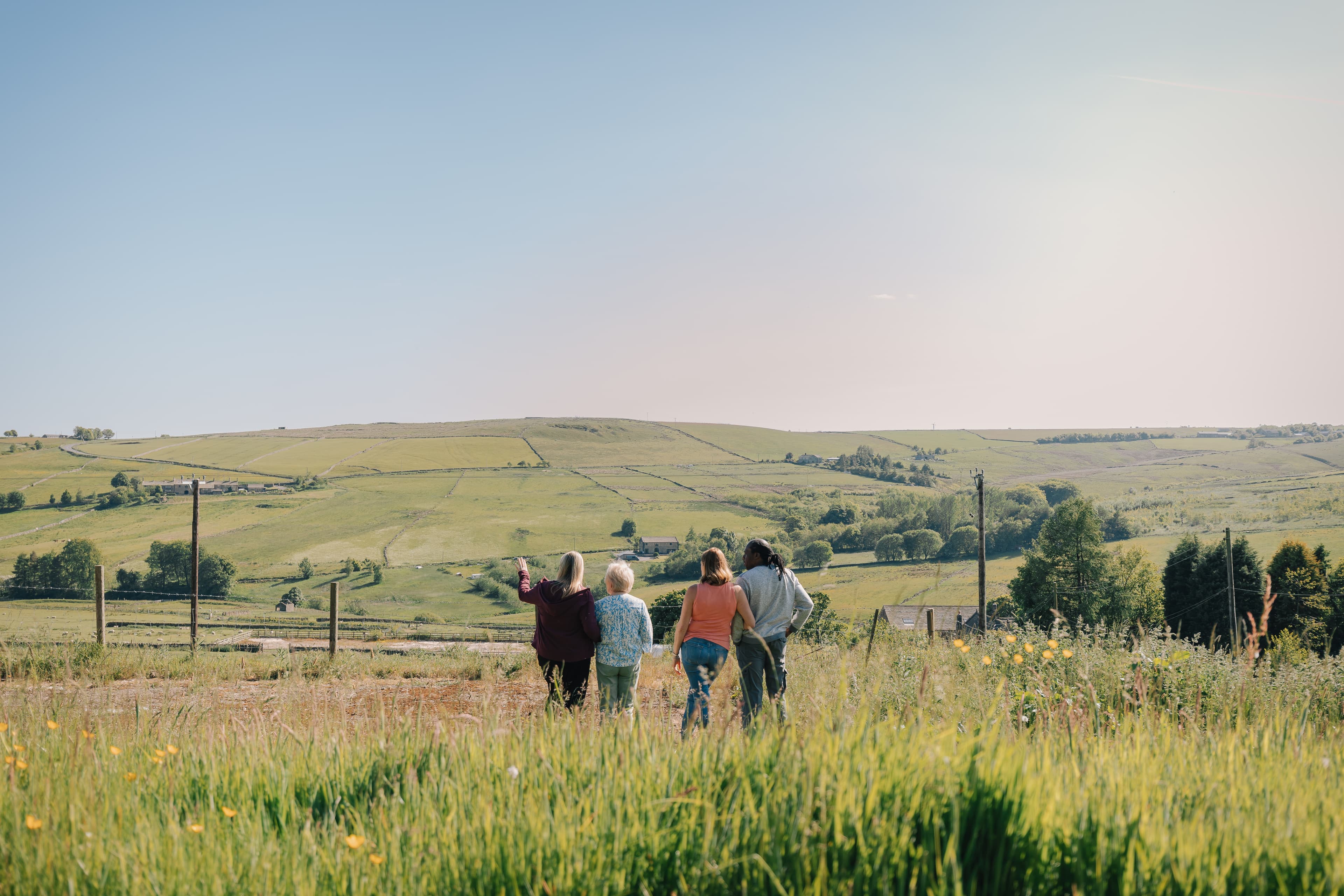 Four people stand in a grassy field, looking out over rolling green hills under a clear sky. - Home Instead