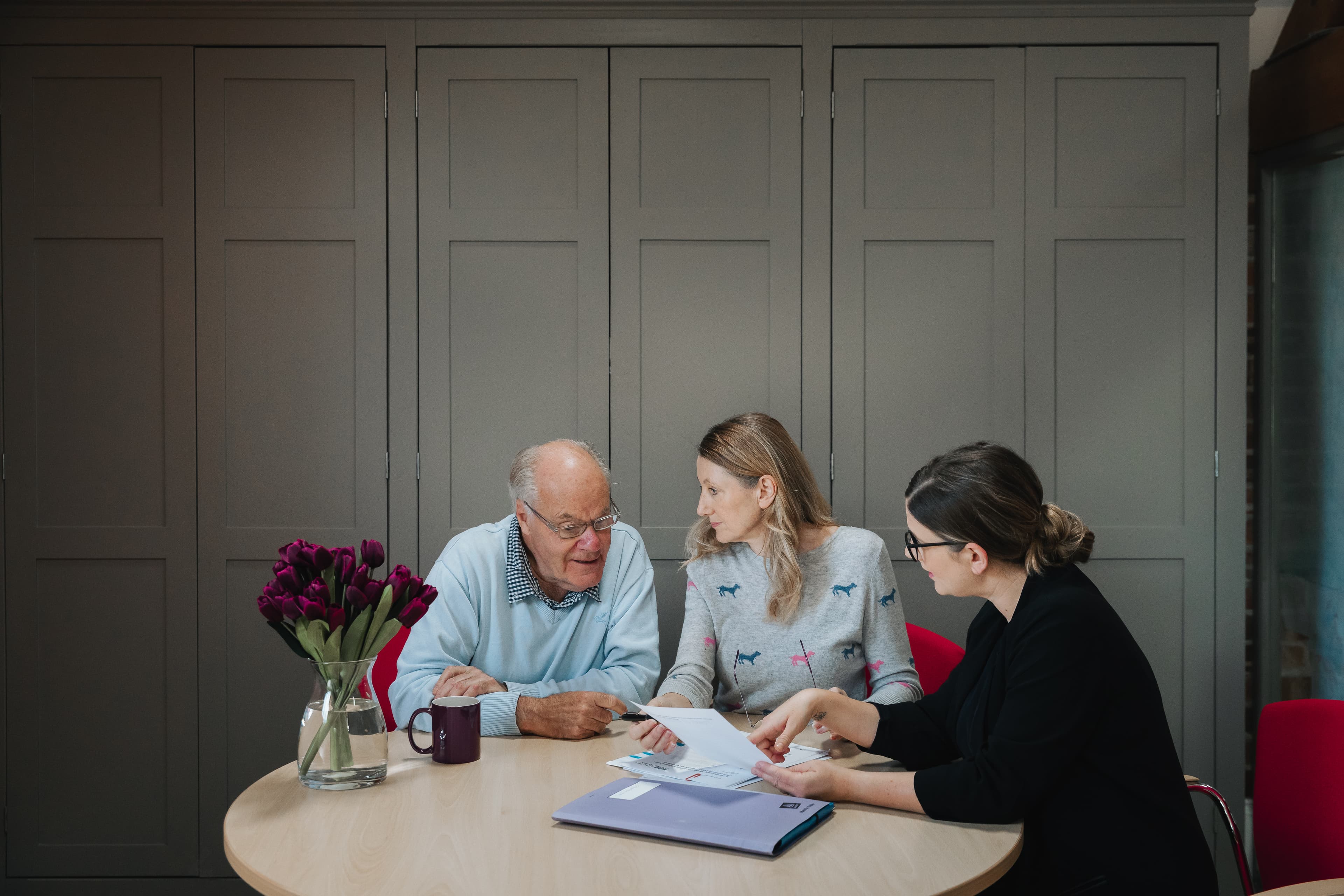 Three people sit at a round table discussing papers, with a vase of tulips and a folder in front of them. - Home Instead