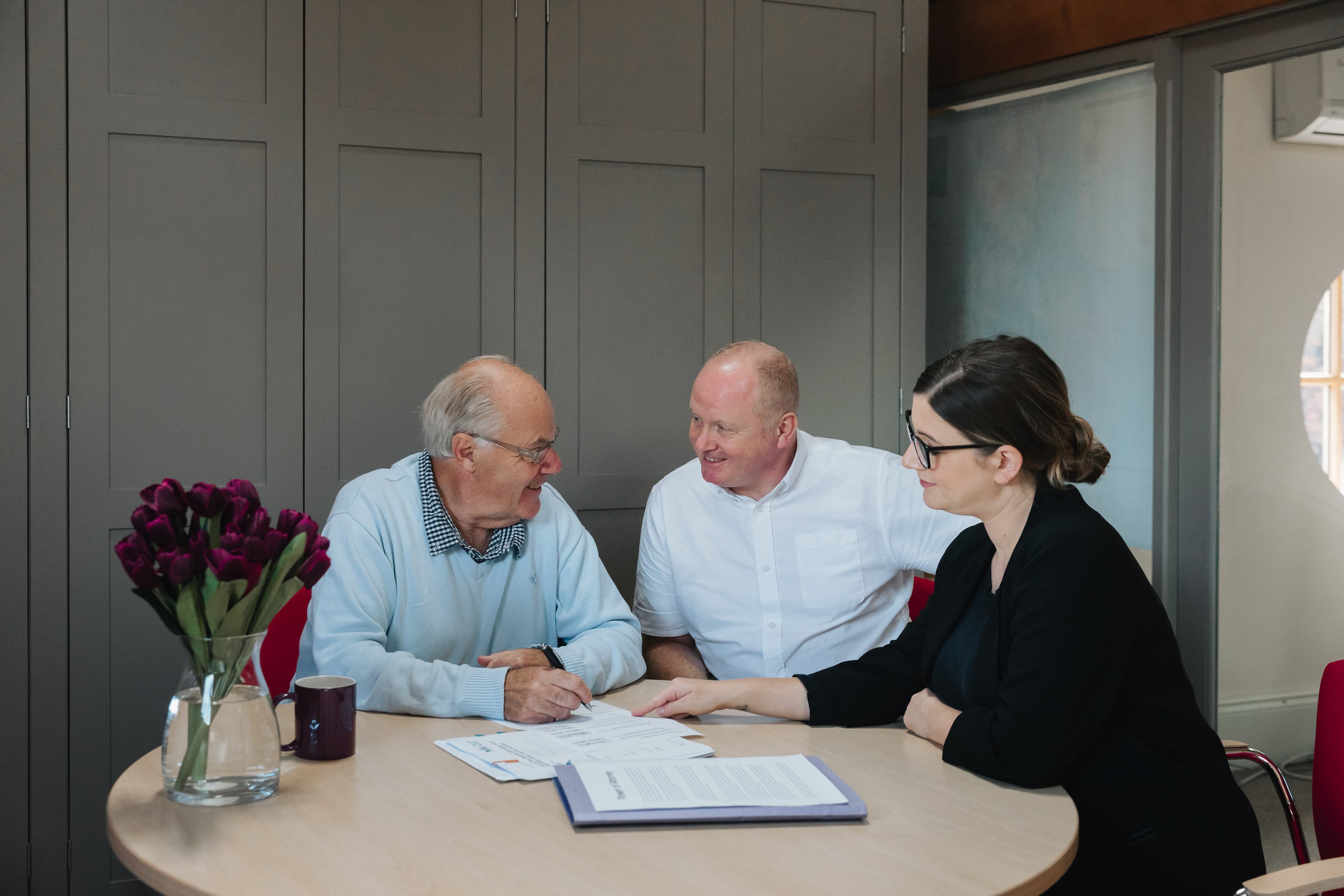 Three people sit at a round table discussing documents, with purple flowers in a vase nearby. - Home Instead