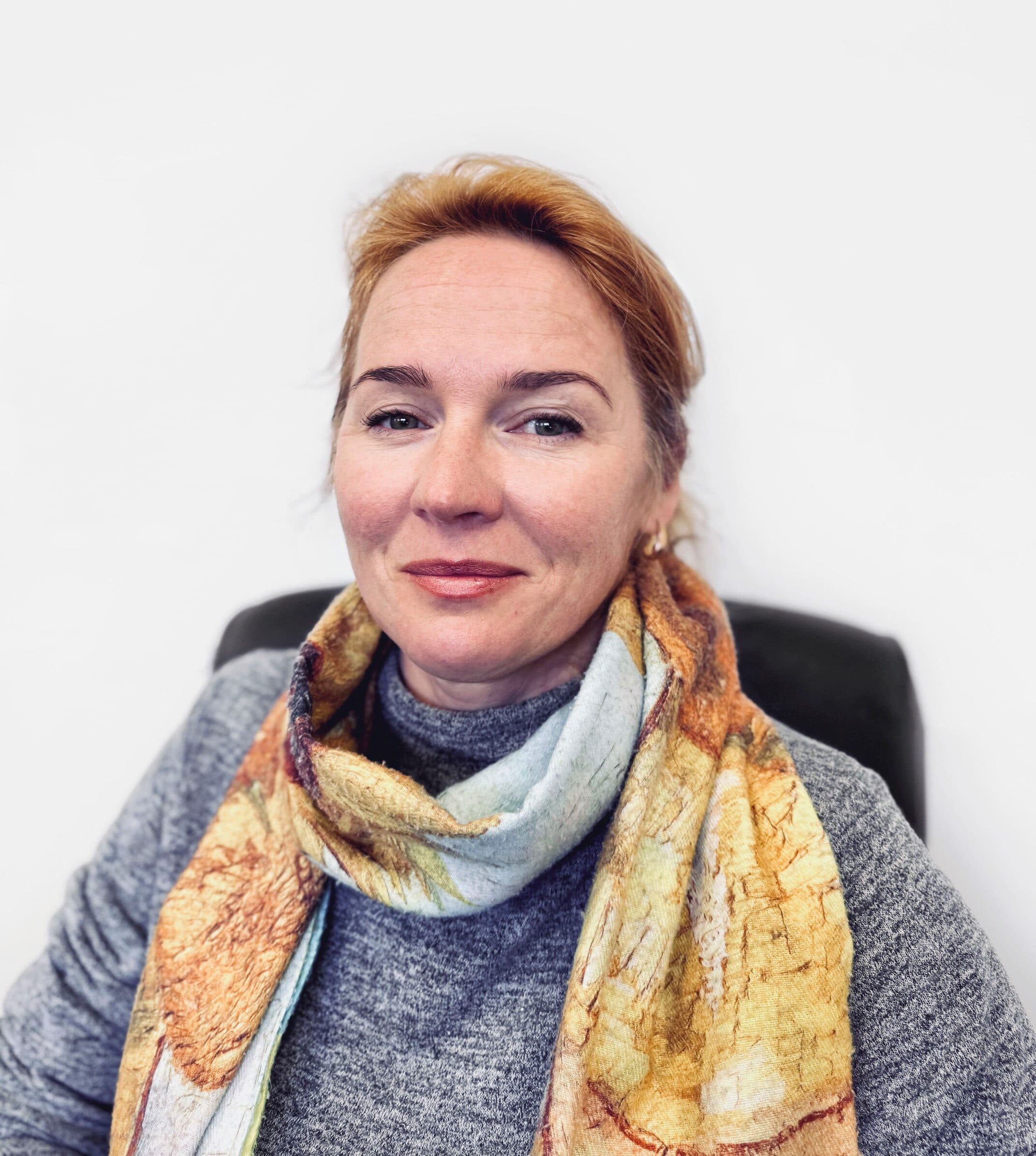 Woman with light brown hair in a gray sweater and colorful scarf, sitting and smiling at the camera. - Home Instead