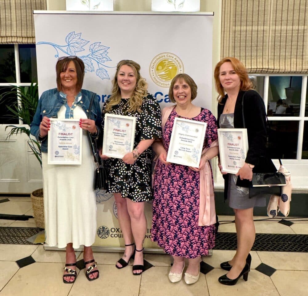 Four women dressed formally stand holding finalist certificates at an awards event in front of a banner. - Home Instead