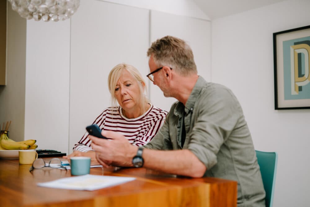 Older couple sit at a table, looking concerned while reviewing information on a smartphone together. - Home Instead