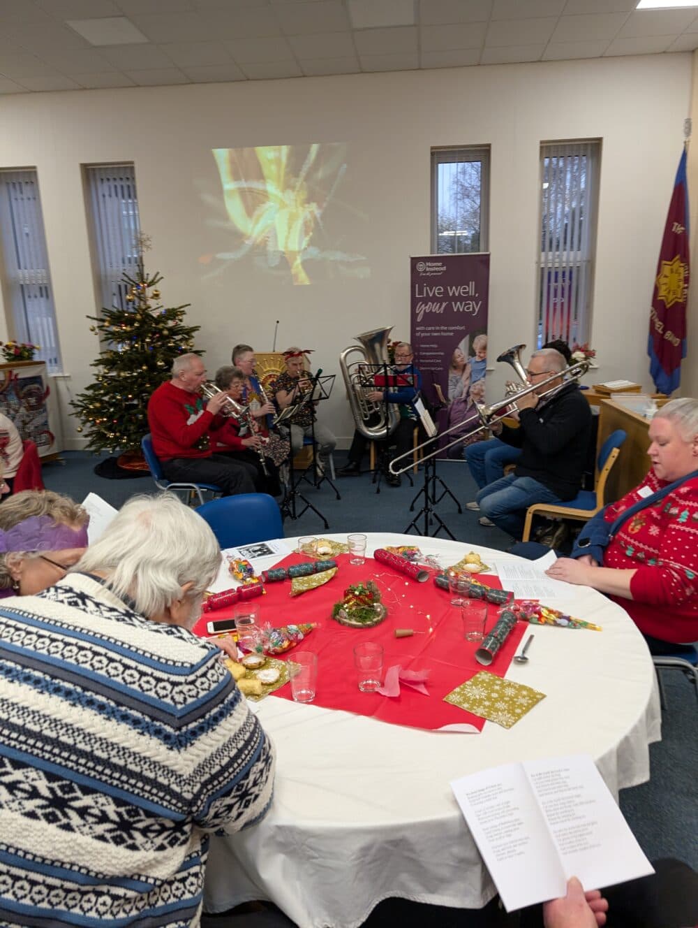 A small brass band plays near a decorated Christmas tree as people sit around a festive table. - Home Instead