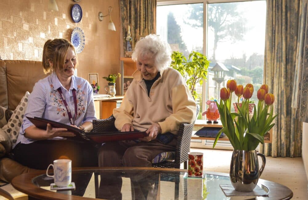 A caregiver and elderly woman smile while looking at a photo album in a cozy living room with flowers on the table. - Home Instead