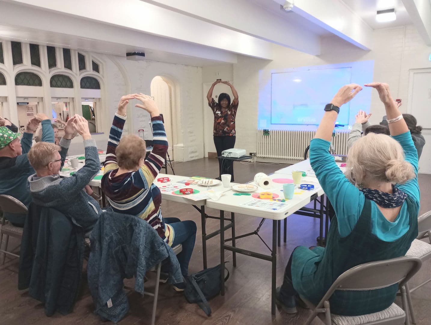 A group of older adults sit around tables, raising their arms in a guided exercise led by an instructor. - Home Instead