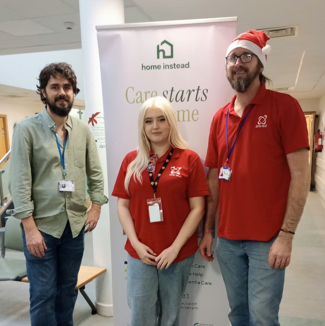 Three people stand in front of a “Home Instead” care banner; one wears a Santa hat and two wear red shirts. - Home Instead