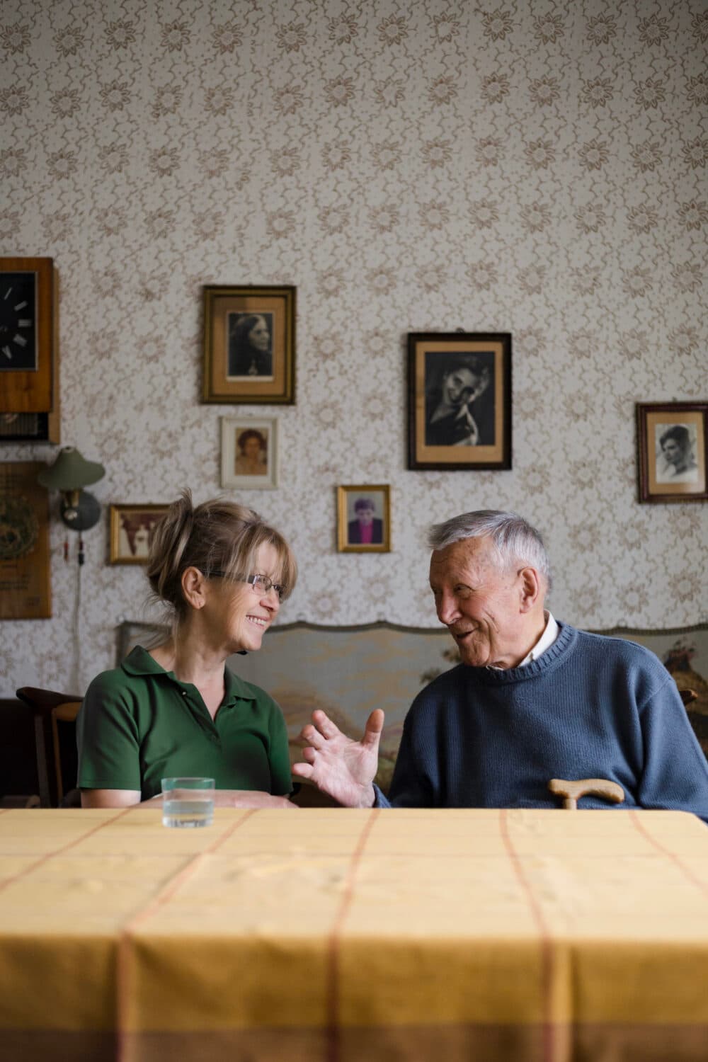 An elderly man and a woman smile and talk at a table in a room with framed photos on the wall. - Home Instead