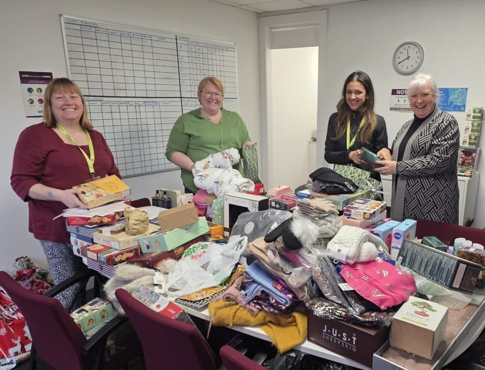 Four smiling women stand around a table covered with assorted donated gifts and supplies in an office. - Home Instead