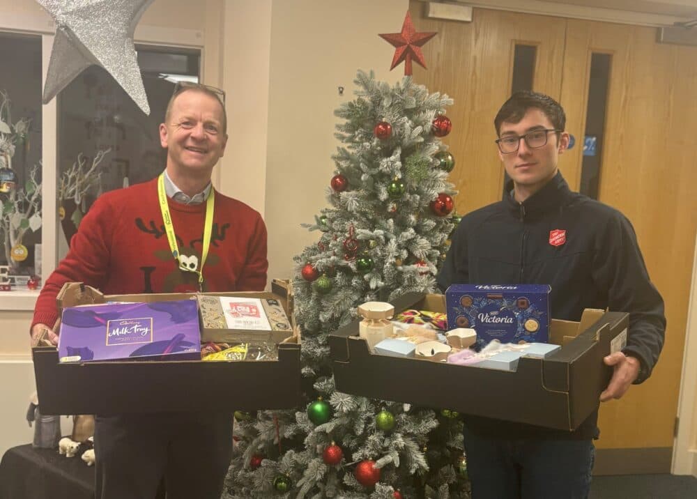 Two men holding gift boxes stand in front of a decorated Christmas tree and festive baubles. - Home Instead