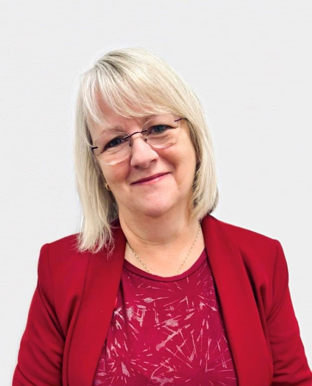 A woman with short blonde hair, glasses, and a red blazer smiles at the camera against a white background. - Home Instead