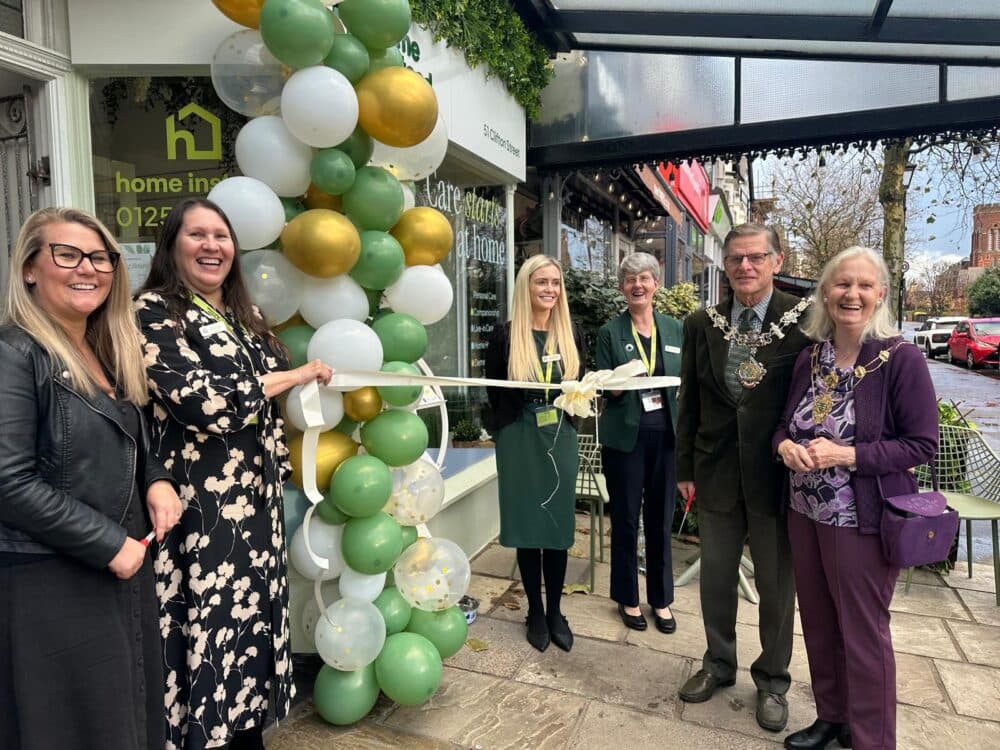 Six people smile at a ribbon-cutting ceremony outside a shop, with green and gold balloons beside them. - Home Instead
