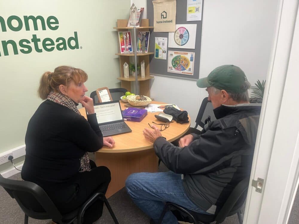 Woman and man sit at a round table in an office, discussing something on a laptop computer. - Home Instead