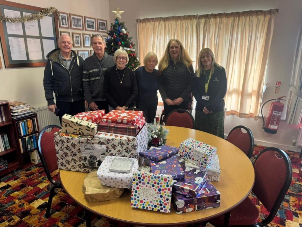 Seven people stand behind a table with wrapped gifts in a festive room with a Christmas tree in the background. - Home Instead