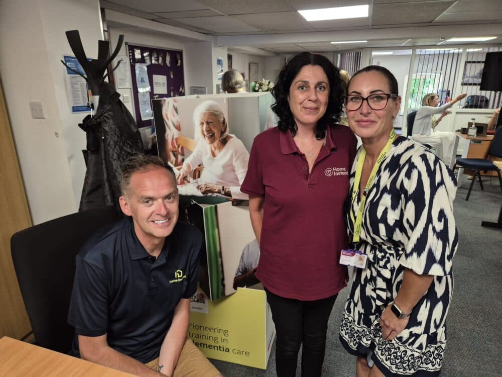 Three adults pose indoors near a dementia care poster, smiling at the camera in an office setting. - Home Instead