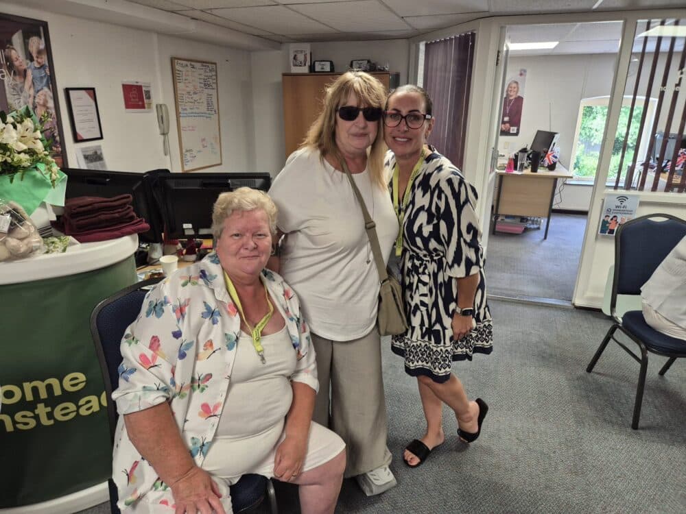 Three women pose and smile together in an office with desks, chairs, and a welcome sign in the background. - Home Instead