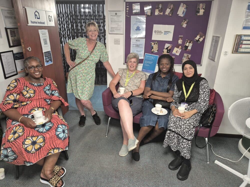 Five women sit and smile with cups of tea in a casual office break area, next to a purple notice board. - Home Instead