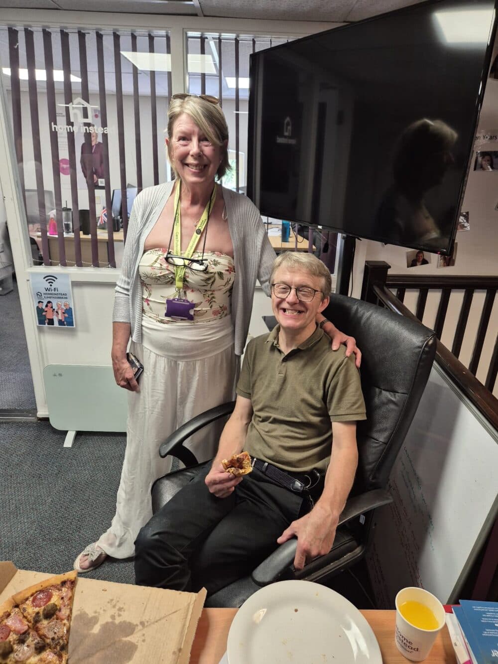 A smiling woman stands by a seated man eating pizza in an office with a TV and pizza box on the table. - Home Instead