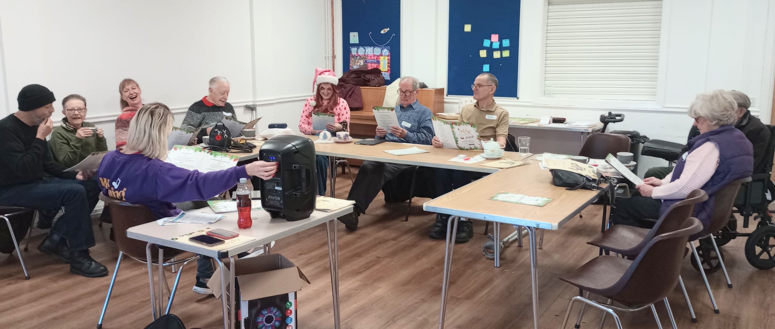 A group of older adults sit around tables in a room, reading papers and laughing together. - Home Instead