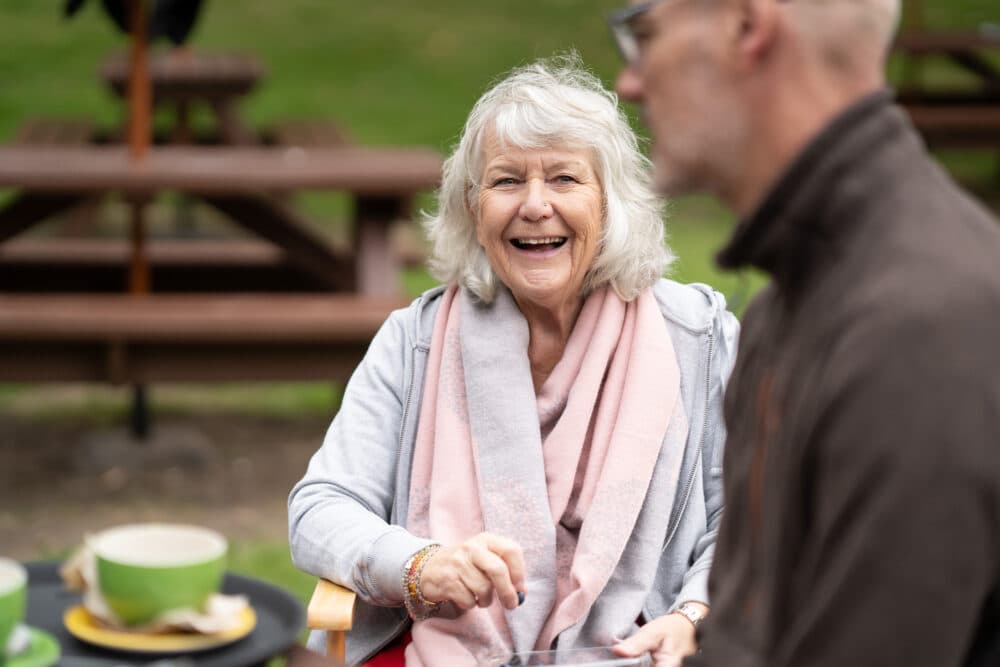 Smiling older woman sitting outdoors at a table, talking to a man in the foreground, with coffee cups nearby. - Home Instead