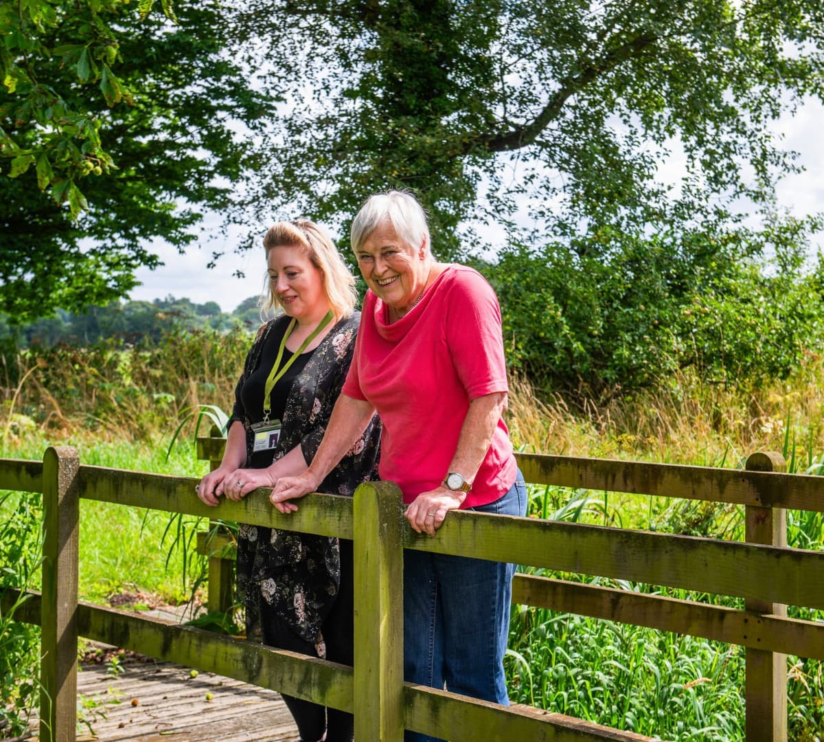 An older female adult with short grey hair and wearing pink standing on a bridge with her younger female carer