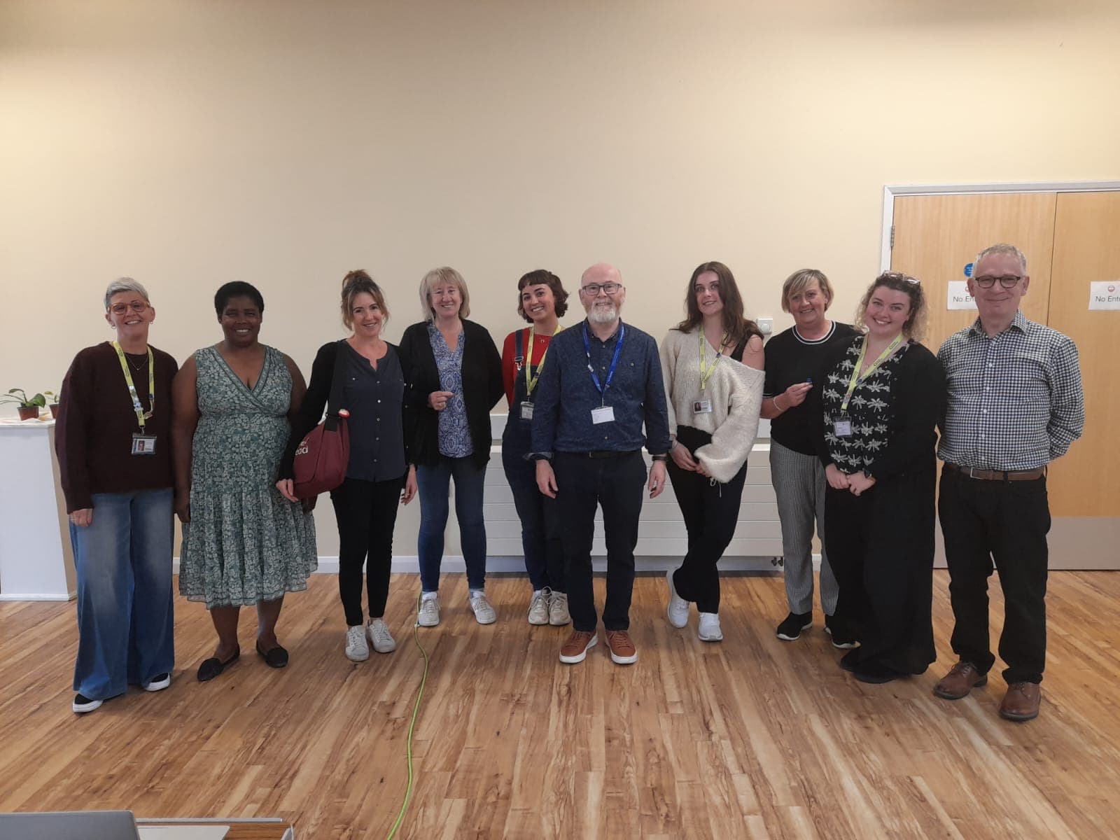 Ten people stand indoors in a row, smiling at the camera in a brightly lit room with wooden flooring. - Home Instead