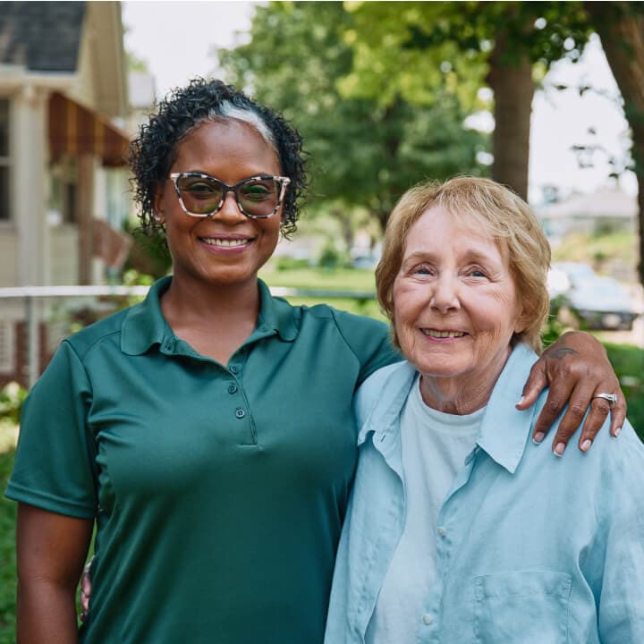 Smiling woman with glasses stands outside with her arm around an older woman on a sunny day. - Home Instead
