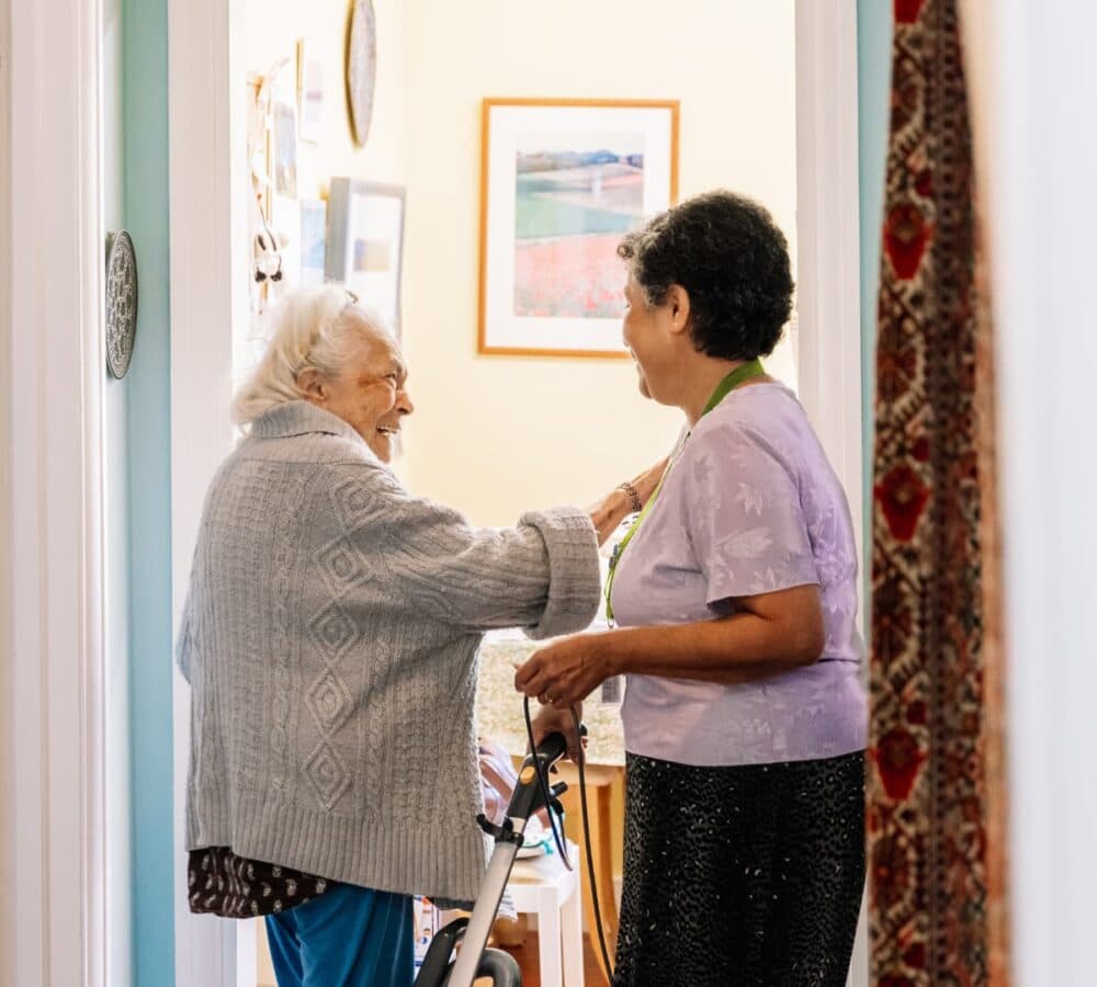 Two elderly women smiling and talking in a corridor, one holding a walking frame and the other touching her shoulder. - Home Instead