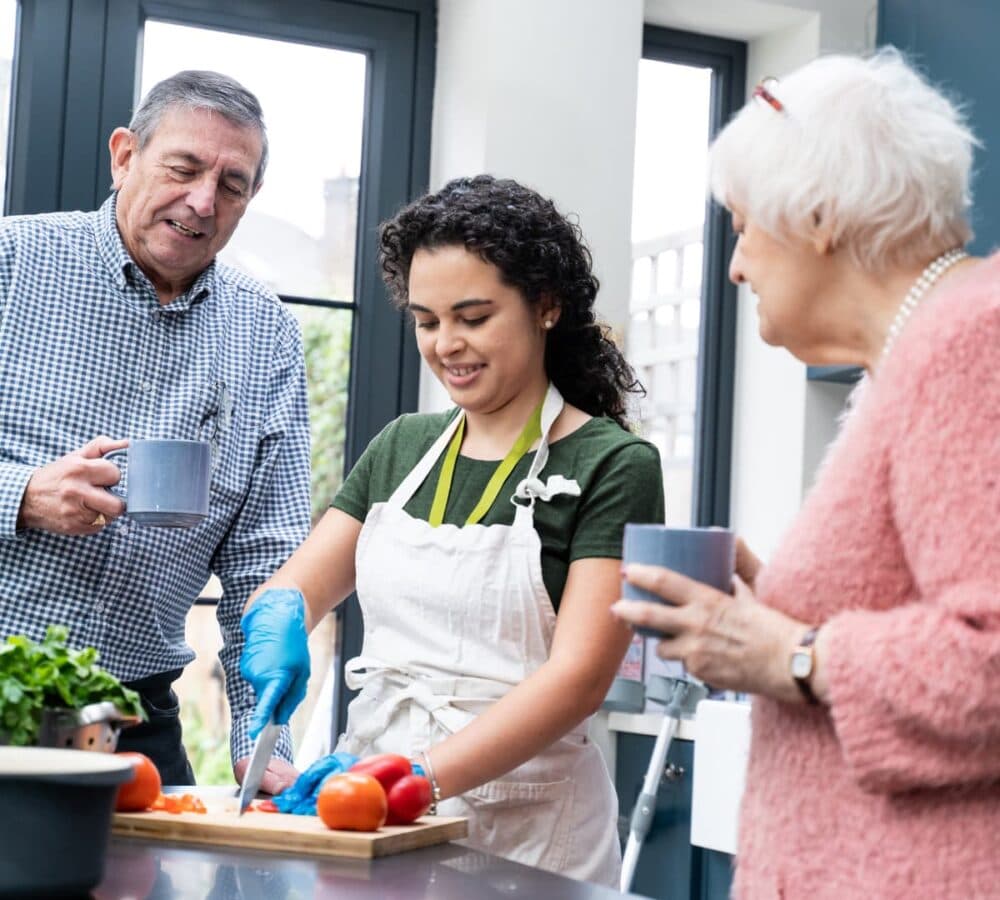 Young woman chopping vegetables while two older adults watch and hold mugs in a bright kitchen. - Home Instead