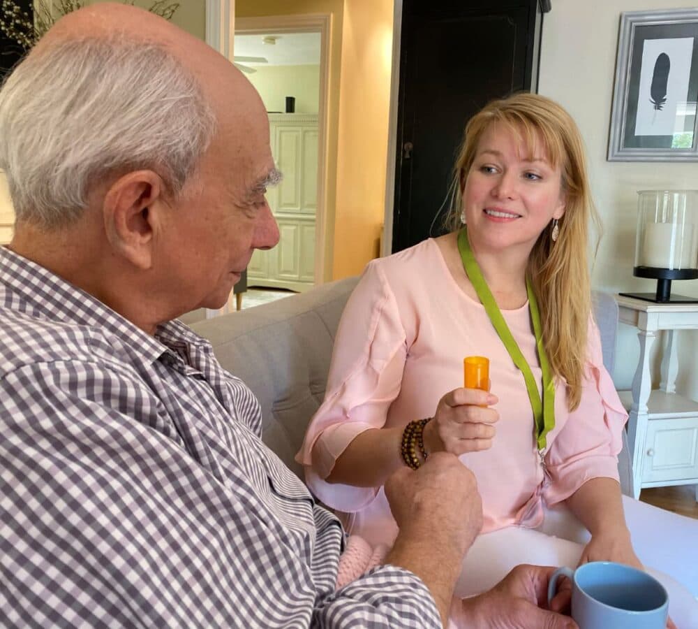 A younger female carer with long blonde hair and wearing pink giving medicines to an older male adult holding a mug and sitting on a couch inside the house