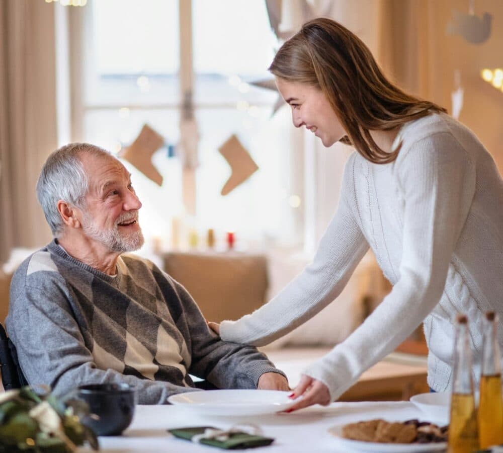 An older male adult with grey hair happy and smiling while sitting on a wheelchair and being helped. y her younger female carer with dark brown hair inside the house