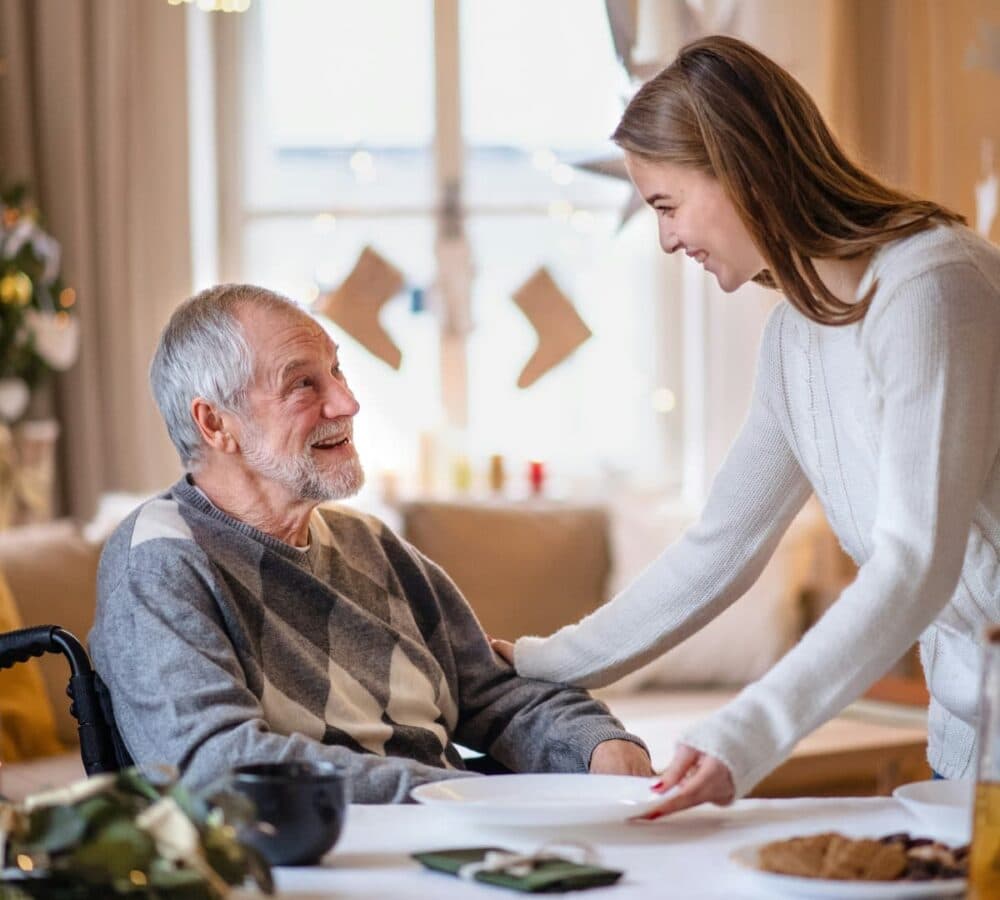 An older male adult with grey hair sitting on a wheel chair happy and smiling with his younger female carer inside the house with christmas tree