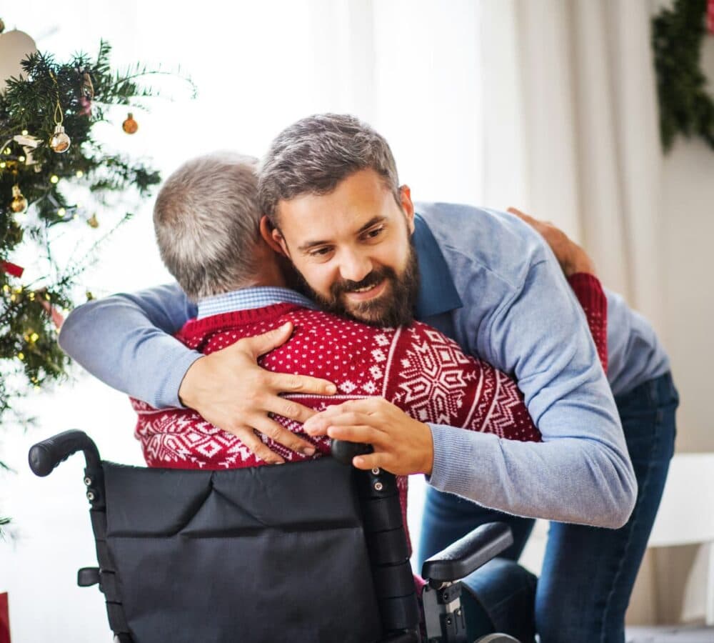 An older male adult sitting on a wheelchair being hugged by a younger male carer with beard