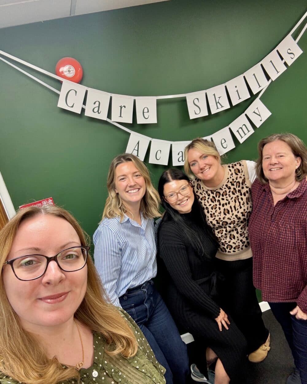 Five women smiling in front of a green wall with a "Care Skills Academy" banner hanging behind them. - Home Instead