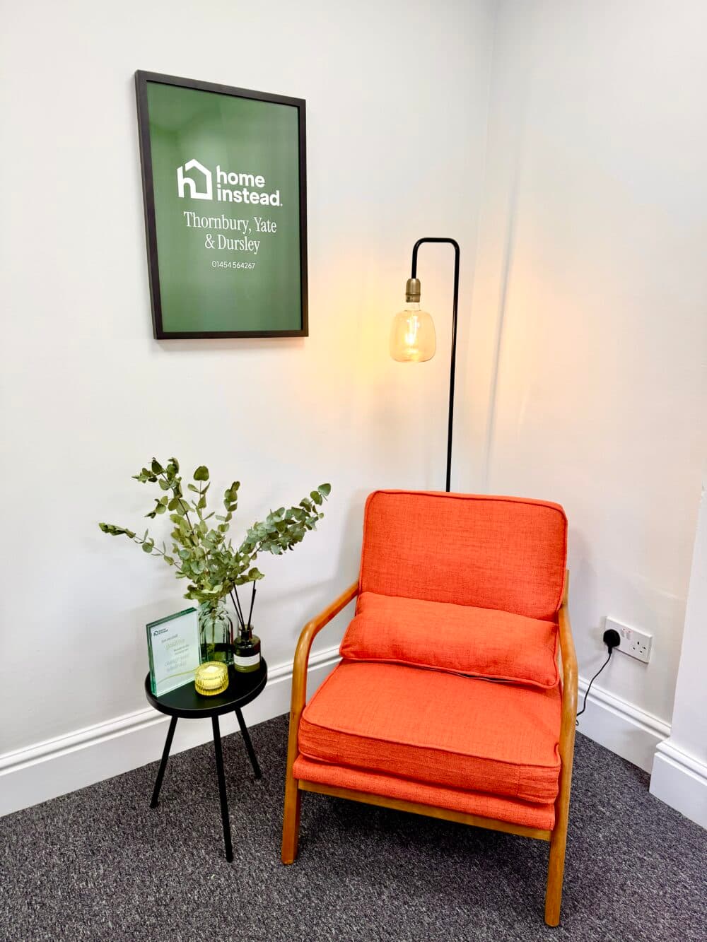 Orange cushioned chair, side table with plants, lamp, and framed sign on a white wall in a cosy corner. - Home Instead
