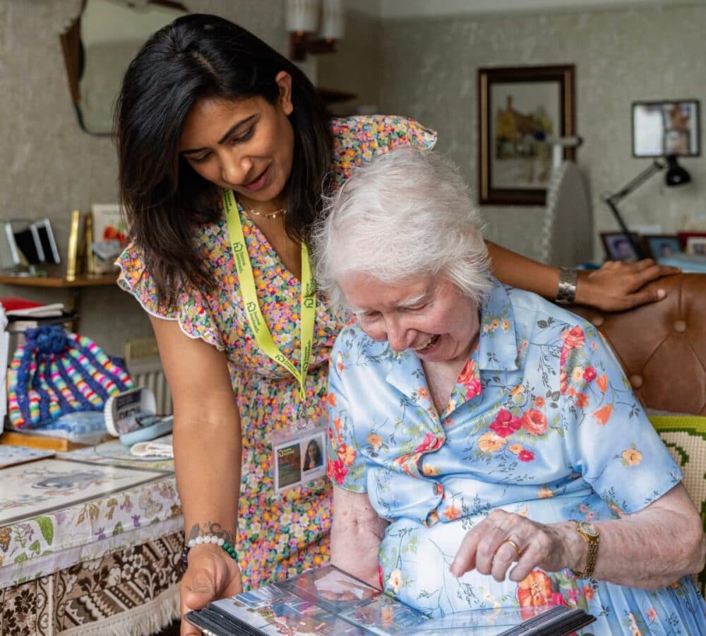 A carer and elderly woman smiling whilst looking at a photo album together in a cosy room. - Home Instead