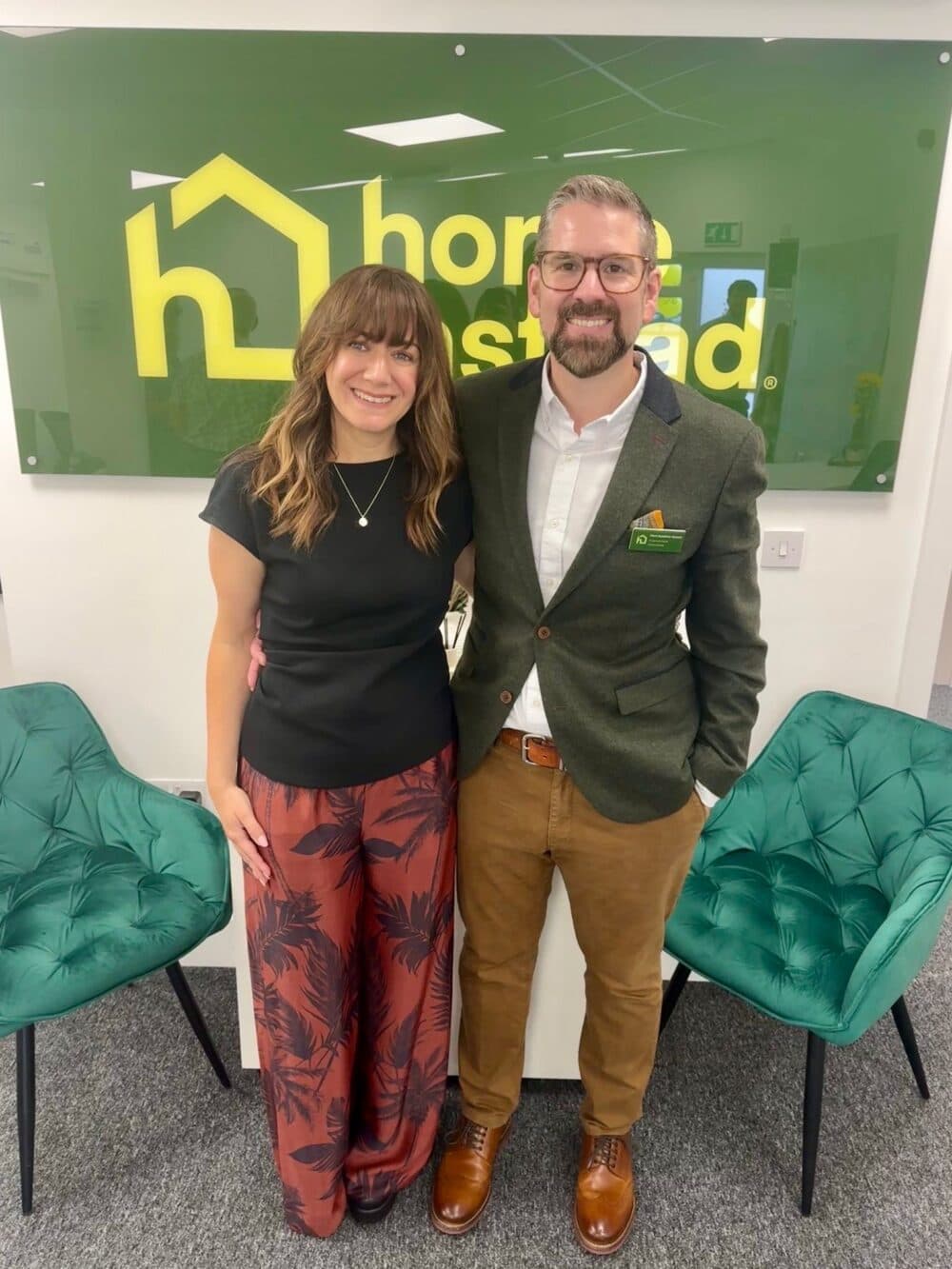Two people standing and smiling in front of a green "homestead" sign, with two green chairs beside them. - Home Instead