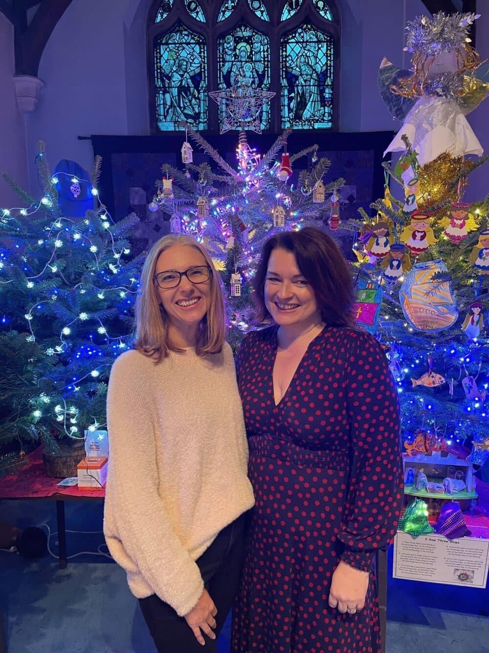 Two women smiling in front of decorated Christmas trees with blue lights and a stained glass window behind them. - Home Instead