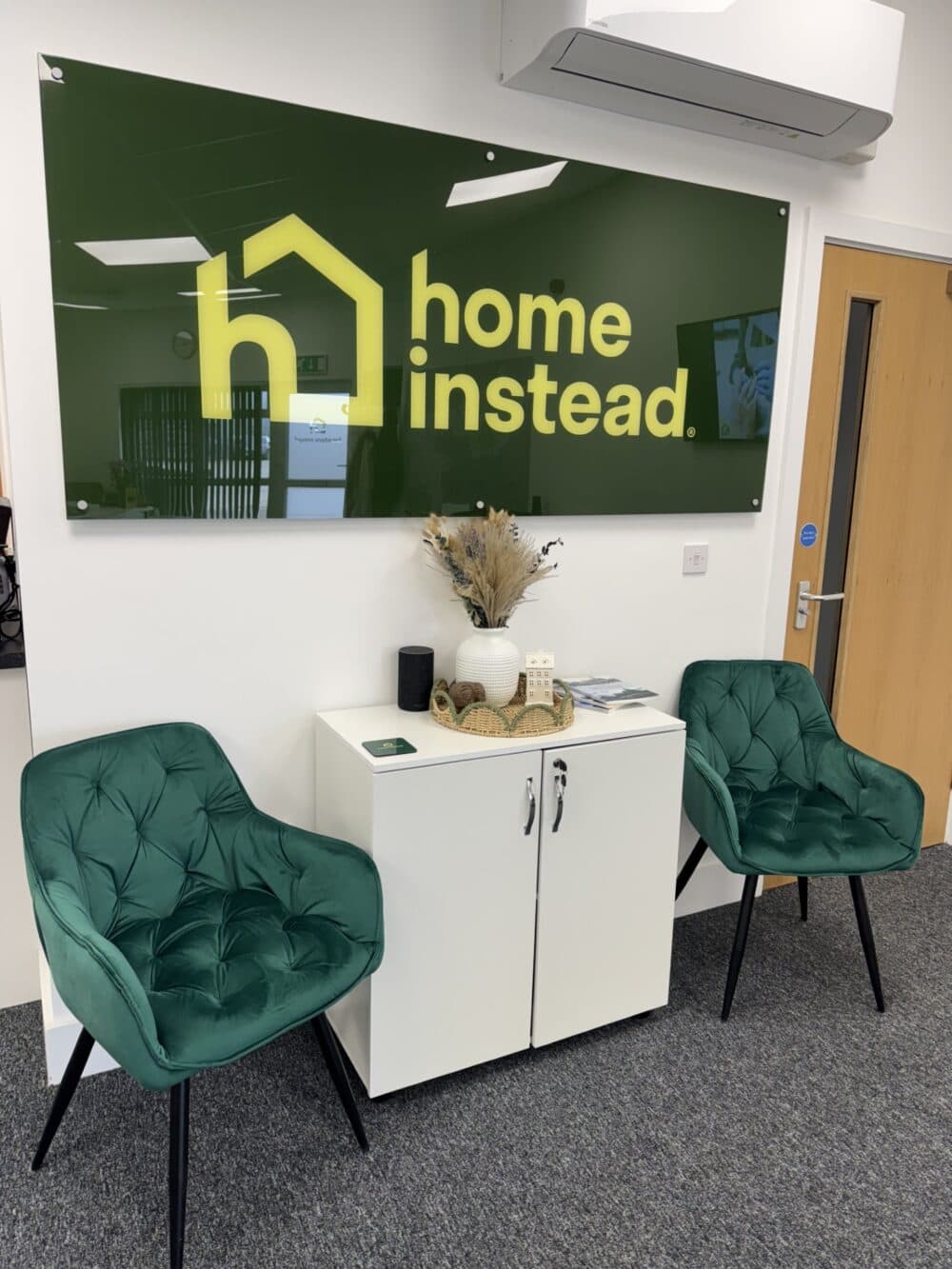 Two green chairs and a white cabinet under a "home instead" sign in a modern office waiting area. - Home Instead
