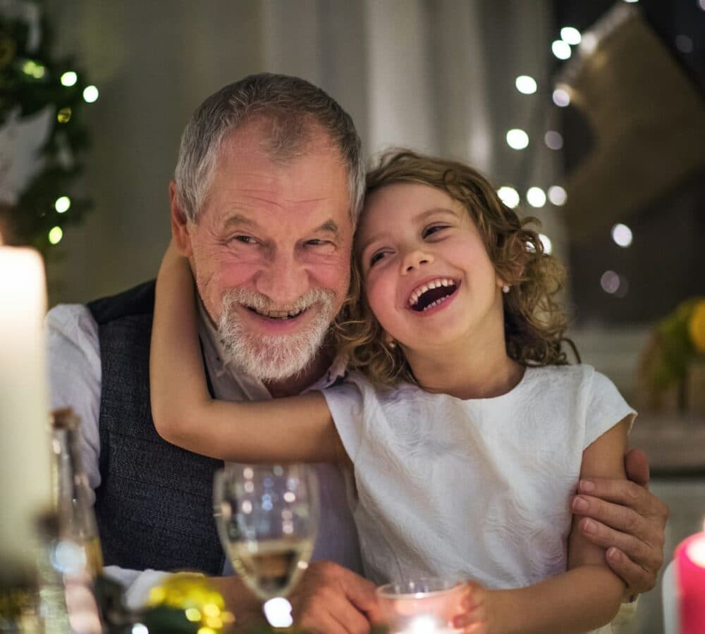 Smiling elderly man and laughing child hug at a festive table with candles and holiday lights in the background. - Home Instead