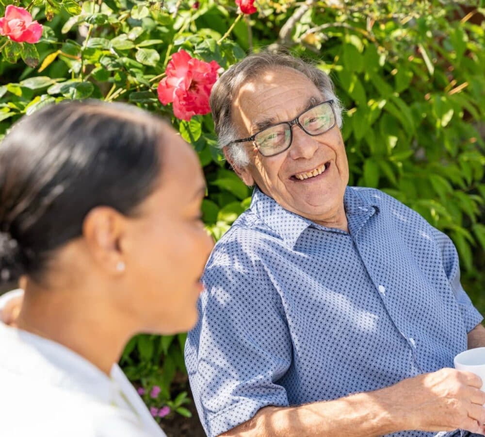 An older male adult wearing eyeglasses and blue polo happy and smiling while sitting in the garden