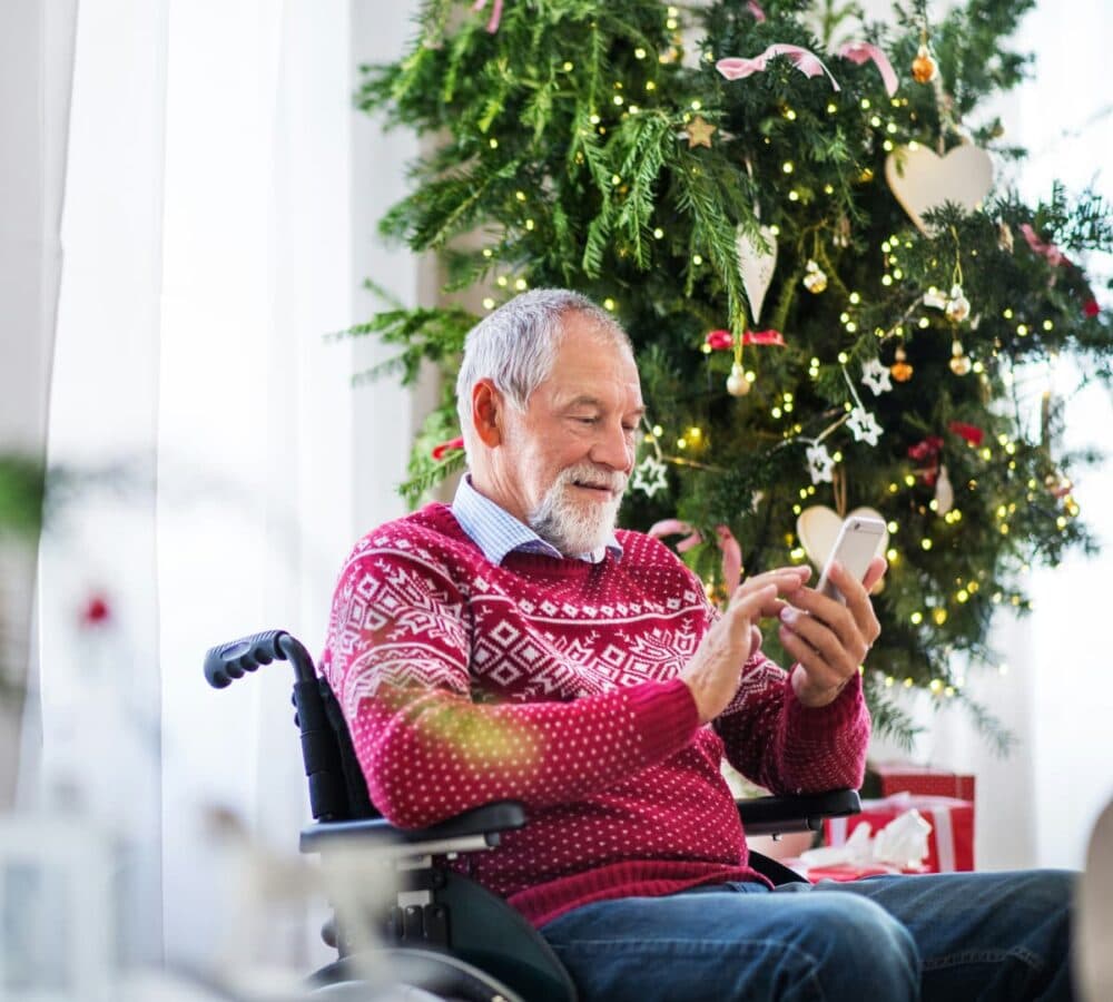 An older male adult with grey hair and beard sitting on a wheelchair and wearing red Christmas sweater while using his cellphone with a Christmas tree beside him inside the house