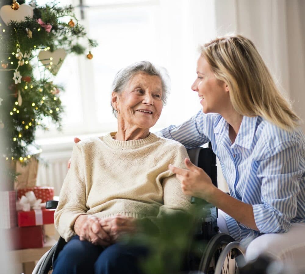 An older female adult with grey hair and witting on a wheelchair happy and smiling while looking at her younger female carer with blonde hair inside the house with christmas tree